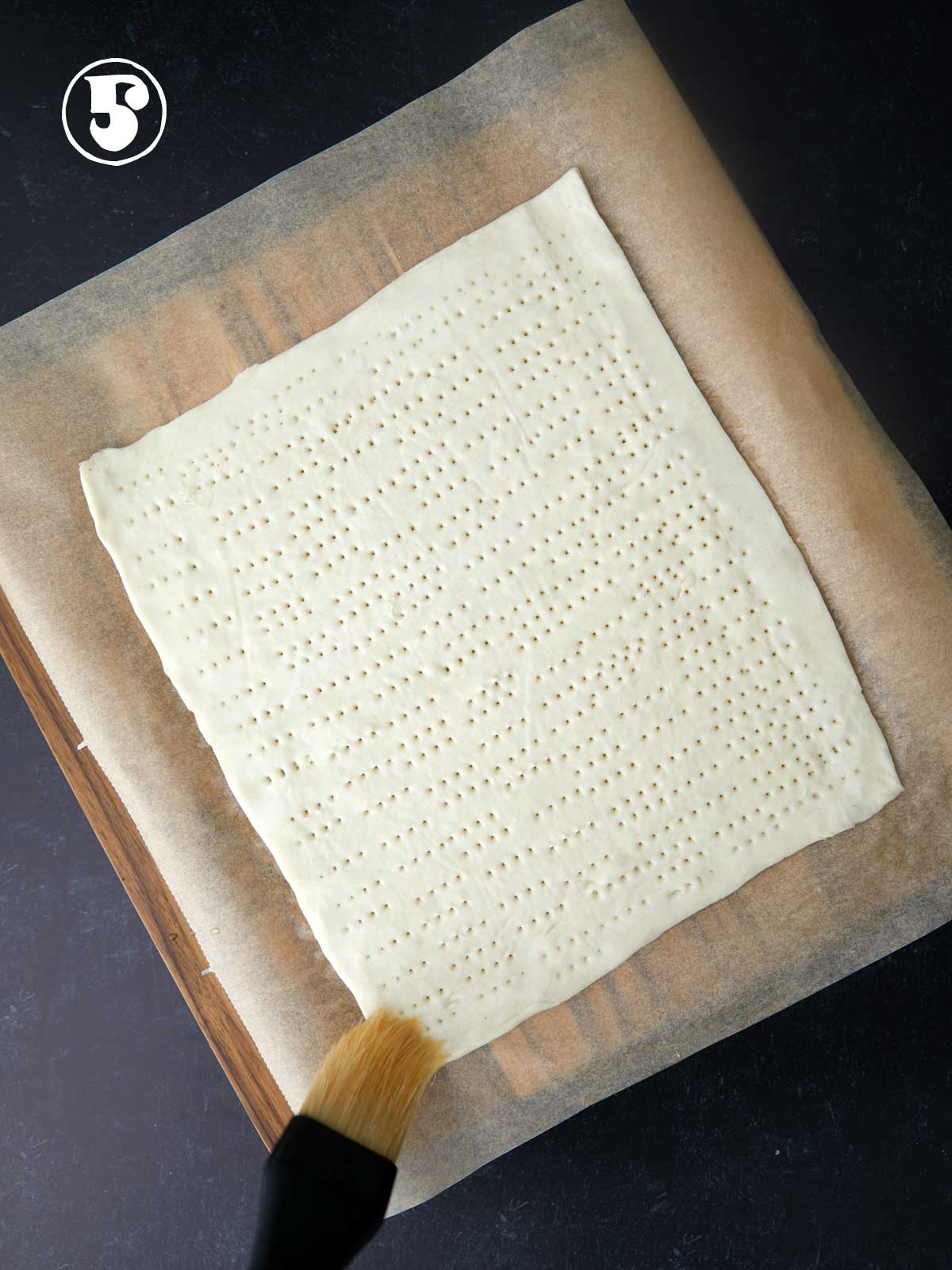 A person is using a brush to apply egg white to the borer of a sheet of puff pastry.