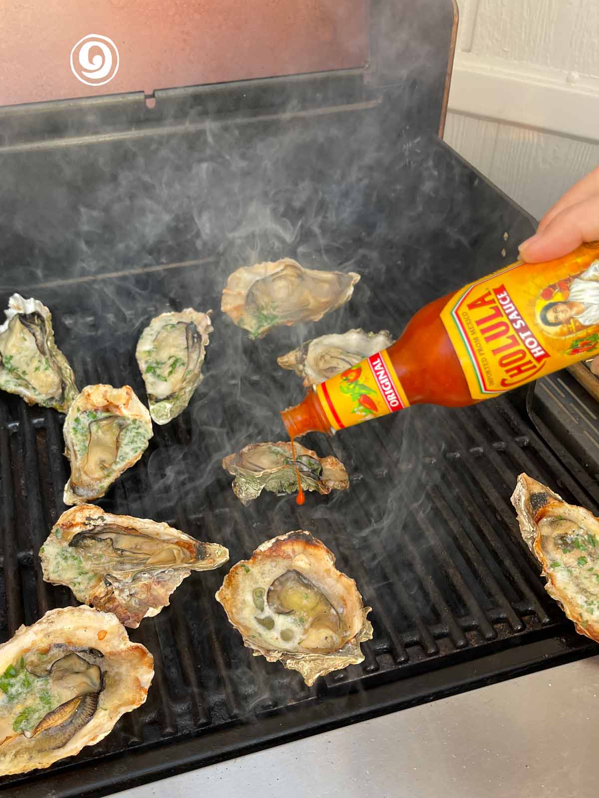 A person adding hot sauce to oysters on the grill.