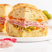 A close-up of a classic Muffuletta sandwich showcasing layers of meat, cheese, and chopped vegetables on a round bread roll, served on a white plate.