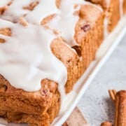 Close-up of frosted apple cake slices on a white tray, accompanied by two cinnamon sticks on a plaid cloth in the foreground, evoking the warm aroma of fresh apple coffee cake.