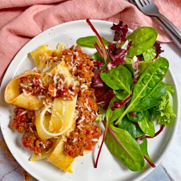 A plate of pappardelle pasta, bolognese sauce, and a small salad.