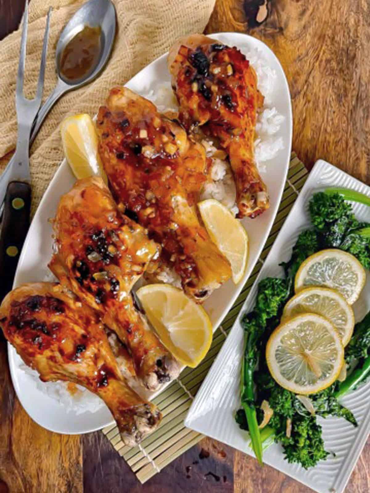 Chicken drumsticks on a serving plate with rice, lemons and broccoli rabe on the side.