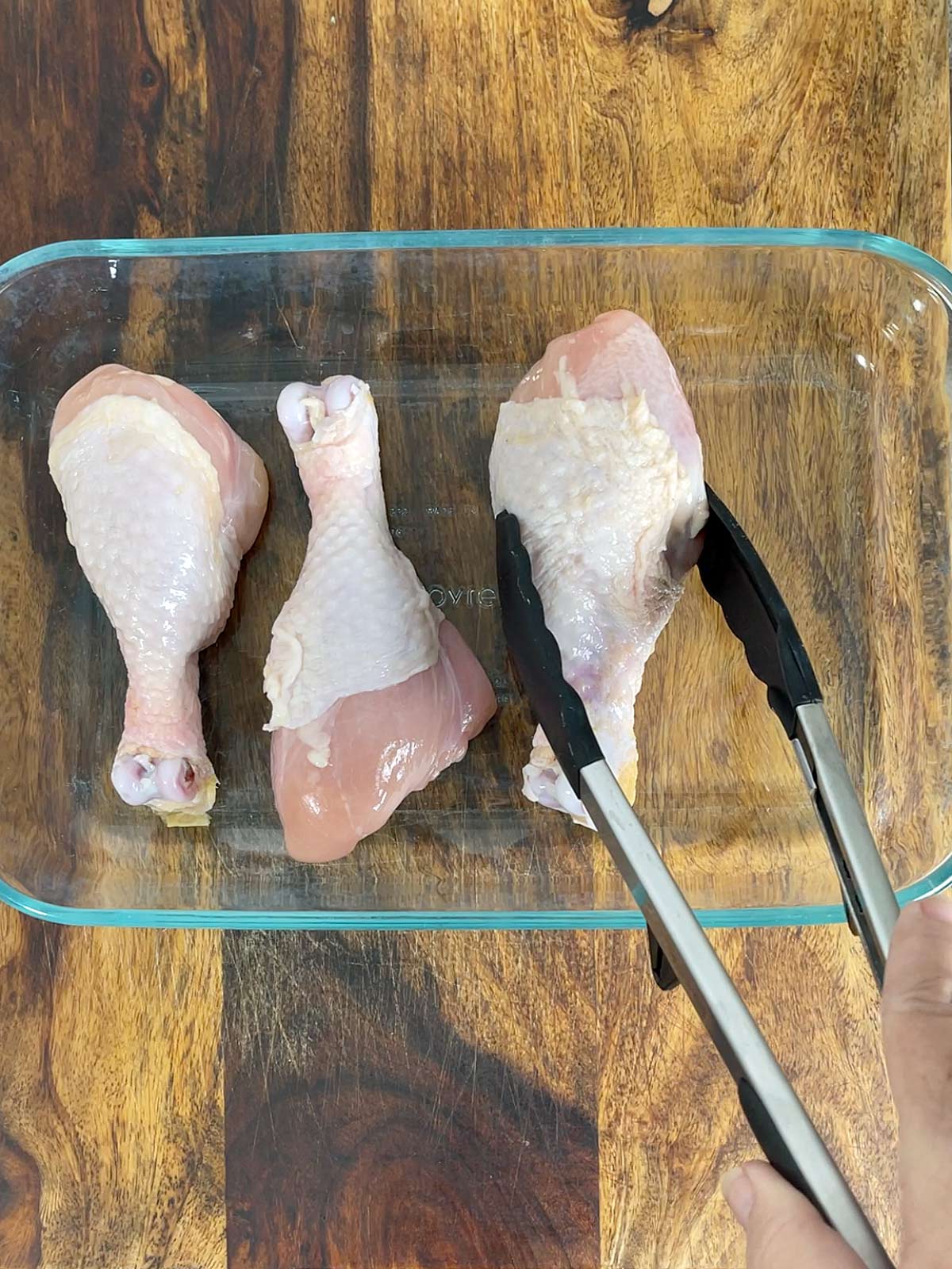 Chicken drumsticks being placed into a glass baking dish with tongs.