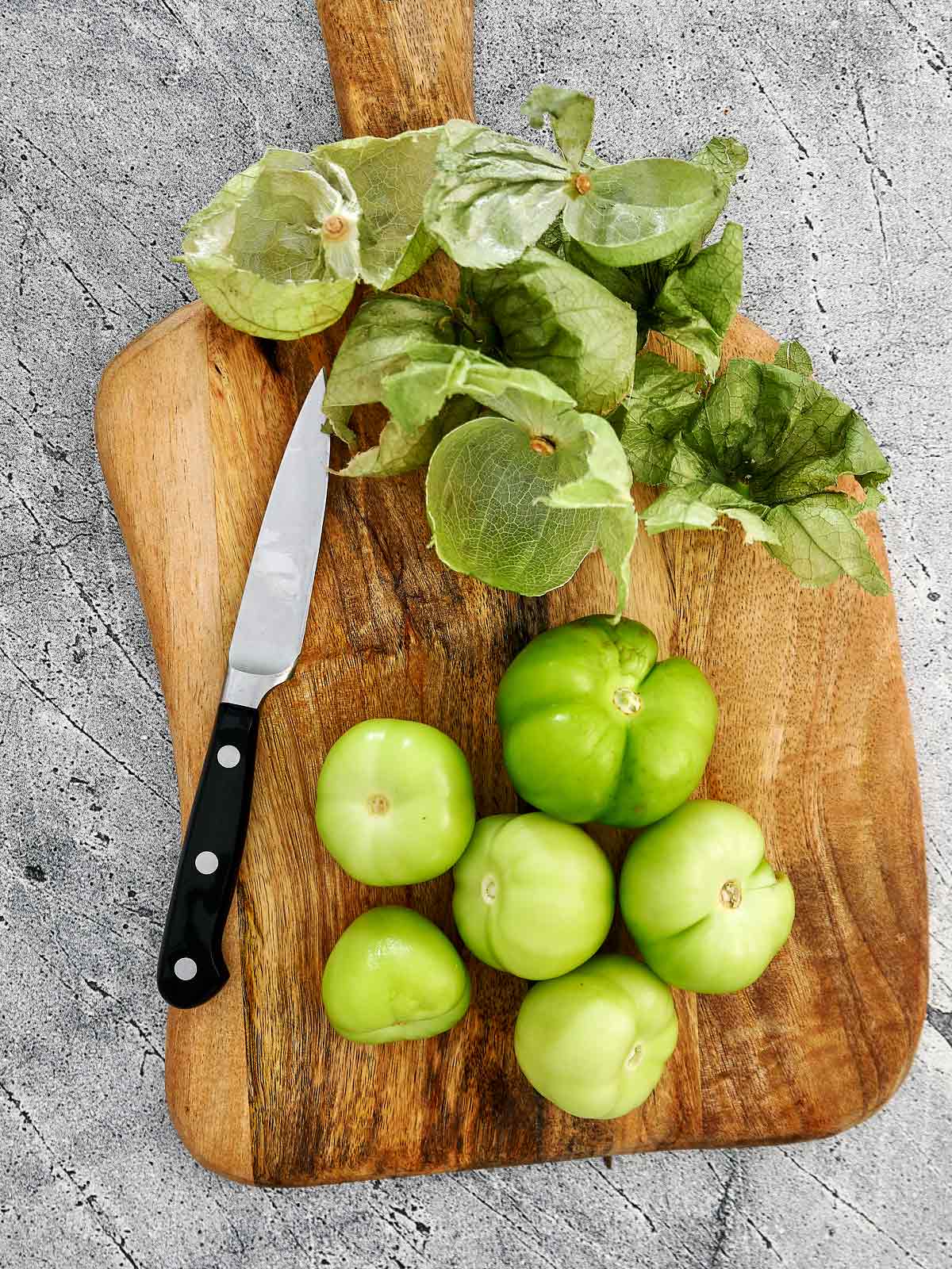 Wooden cutting board with tomatillos and the papery husks that have been removed.