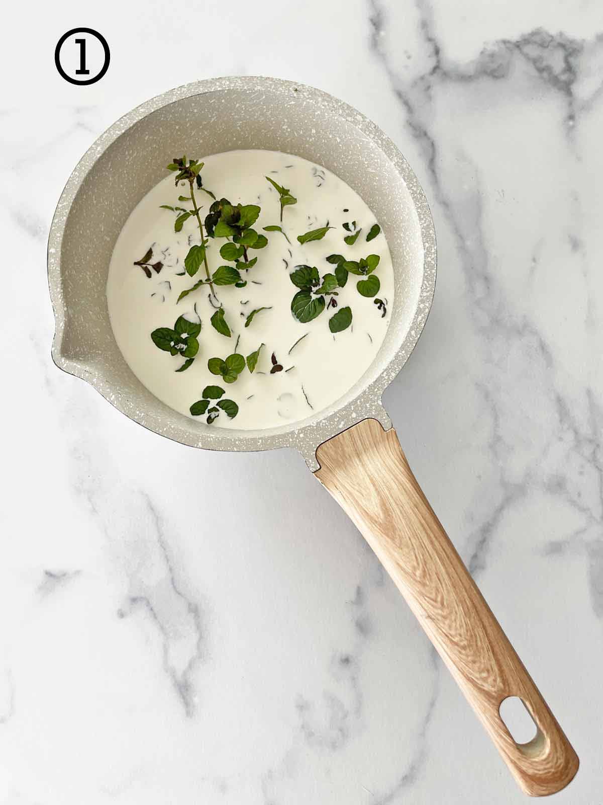 A sauce pan with a wooden handle resting on a marble countertop with milk and mint in it.
