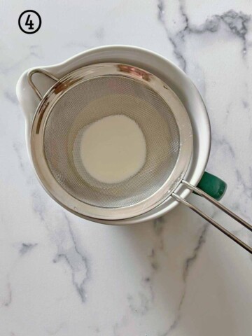 A stainless steel strainer with a handle on it, atop a bowl of cream.