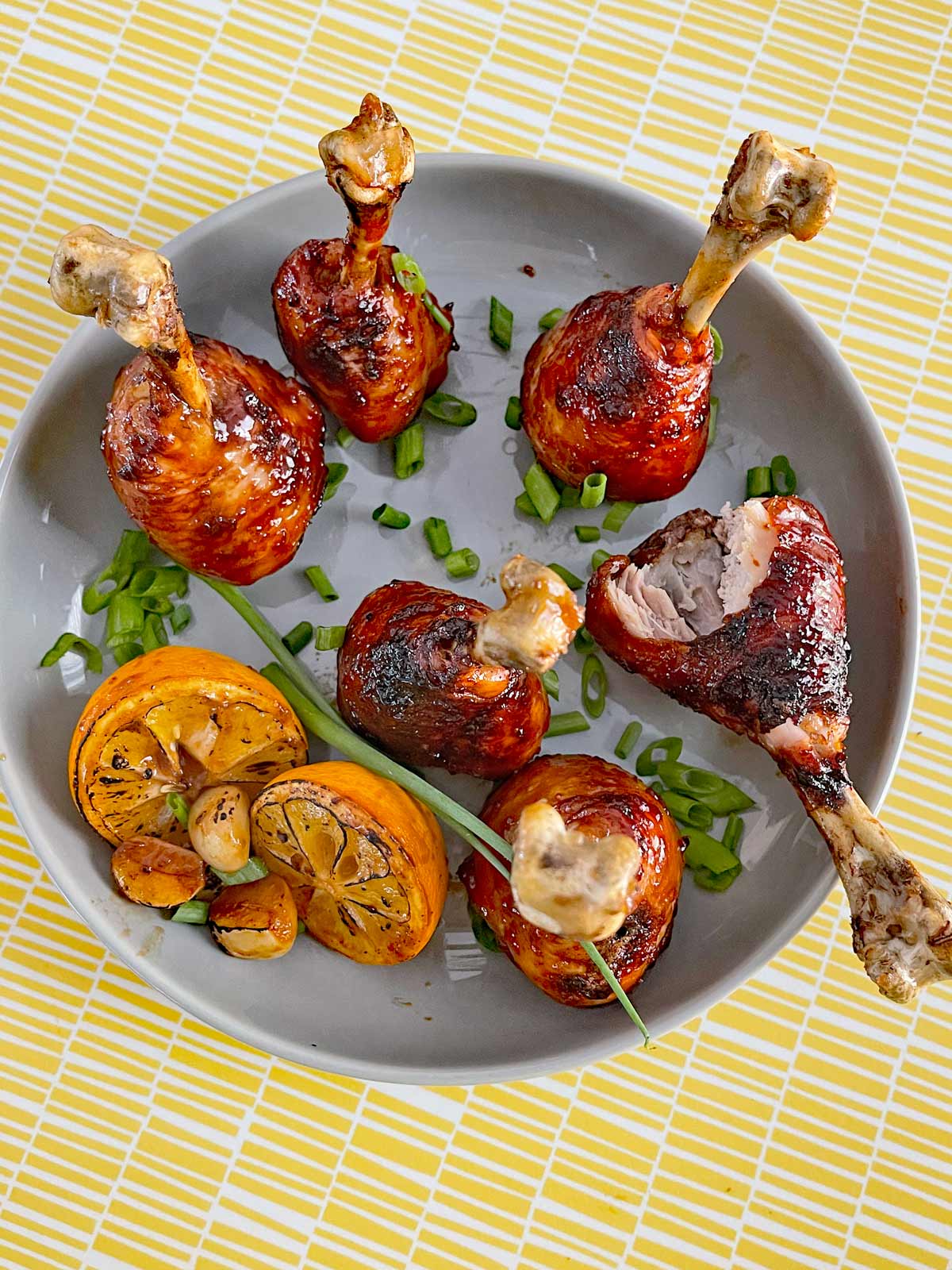 Plated chicken lollipops with lemon and garlic garnish and sprinkled scallions. One drumstick has a big bite missing.