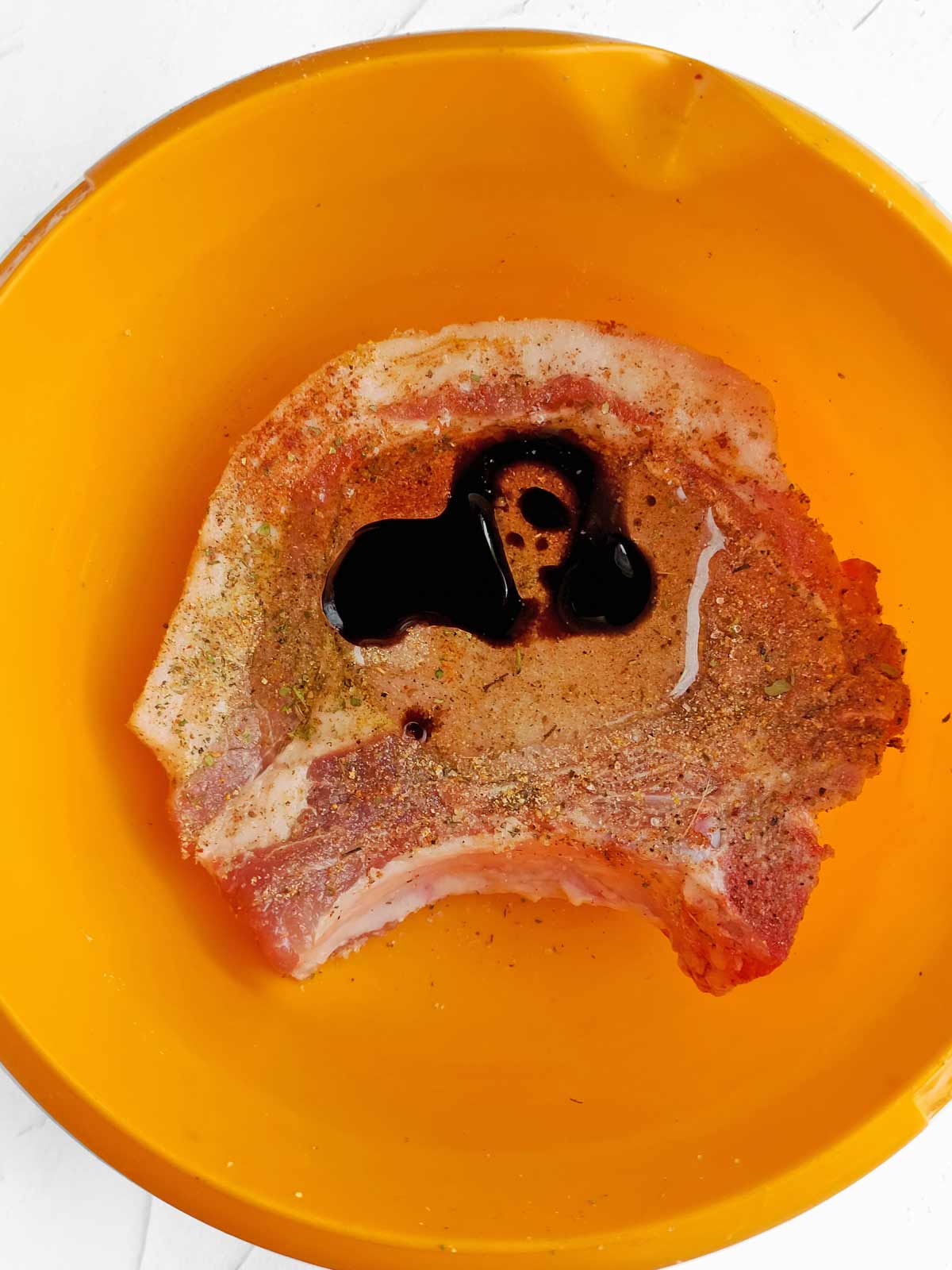 Pork chop in bowl with spices and oil.