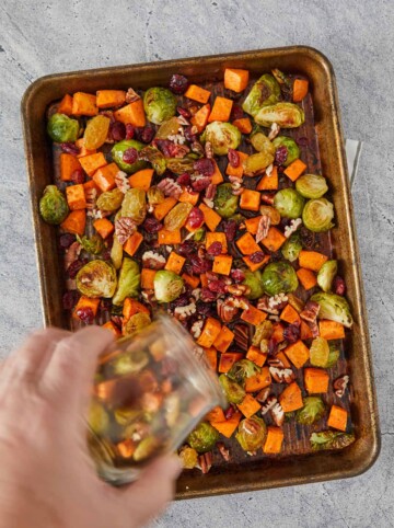 Sheet pan with roasted vegetables, cranberries, pecans, and golden raisins with dressing being poured on from a jar.