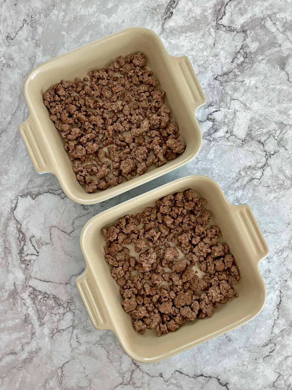 Two small baking dishes with ground beef on the bottoms.