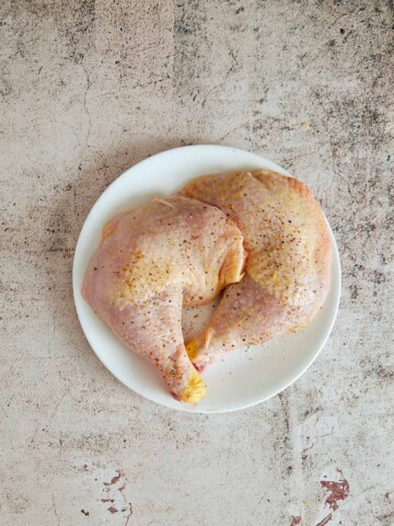 Two pieces of chicken on a plate with salt and pepper sprinkled on.