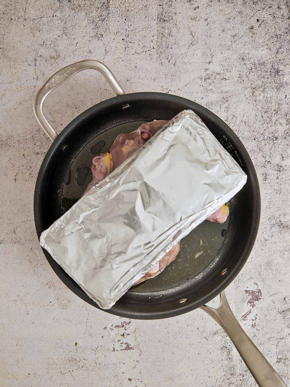 Brick wrapped in foil placed on the flesh side of the chicken pieces in the skillet.