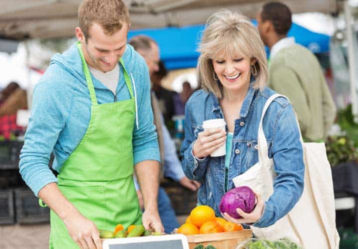 Smiling woman with coffee buying vegetables from farmer's market vendor.