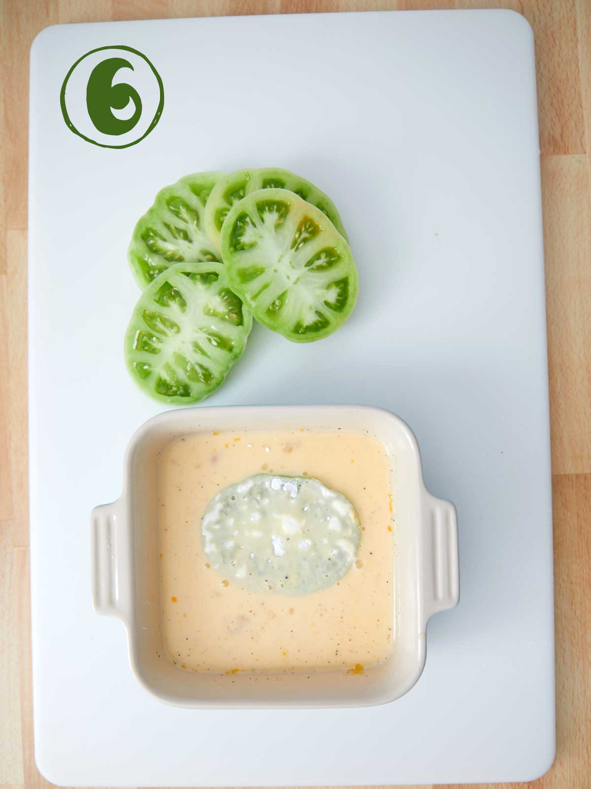 A bowl of egg and buttermilk on a cutting board with green tomatoes.