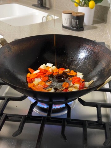 A wok filled with vegetables on a stove top with sauce pouring in.