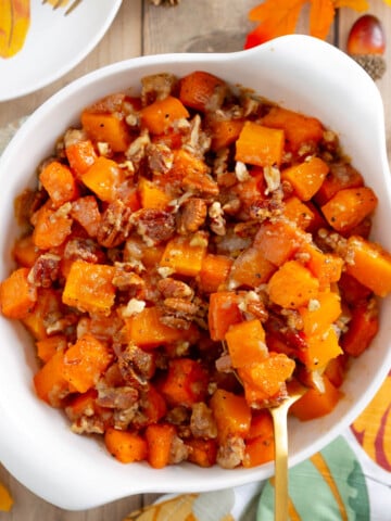 A white bowl filled with roasted butternut squash and sweet potatoes mixed with pecans, served on a surface decorated with autumn leaves.