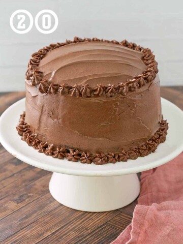 A chocolate cake on a white stand with chocolate buttercream frosting.