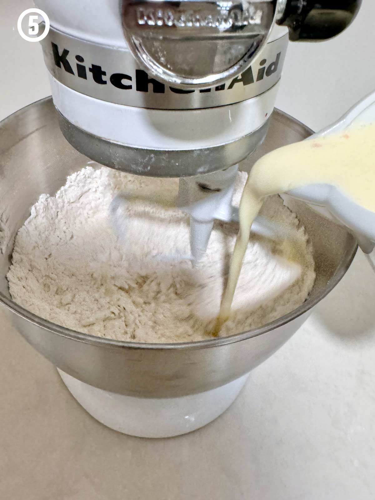 A kitchenaid mixer is pouring milk into a bowl of flour.
