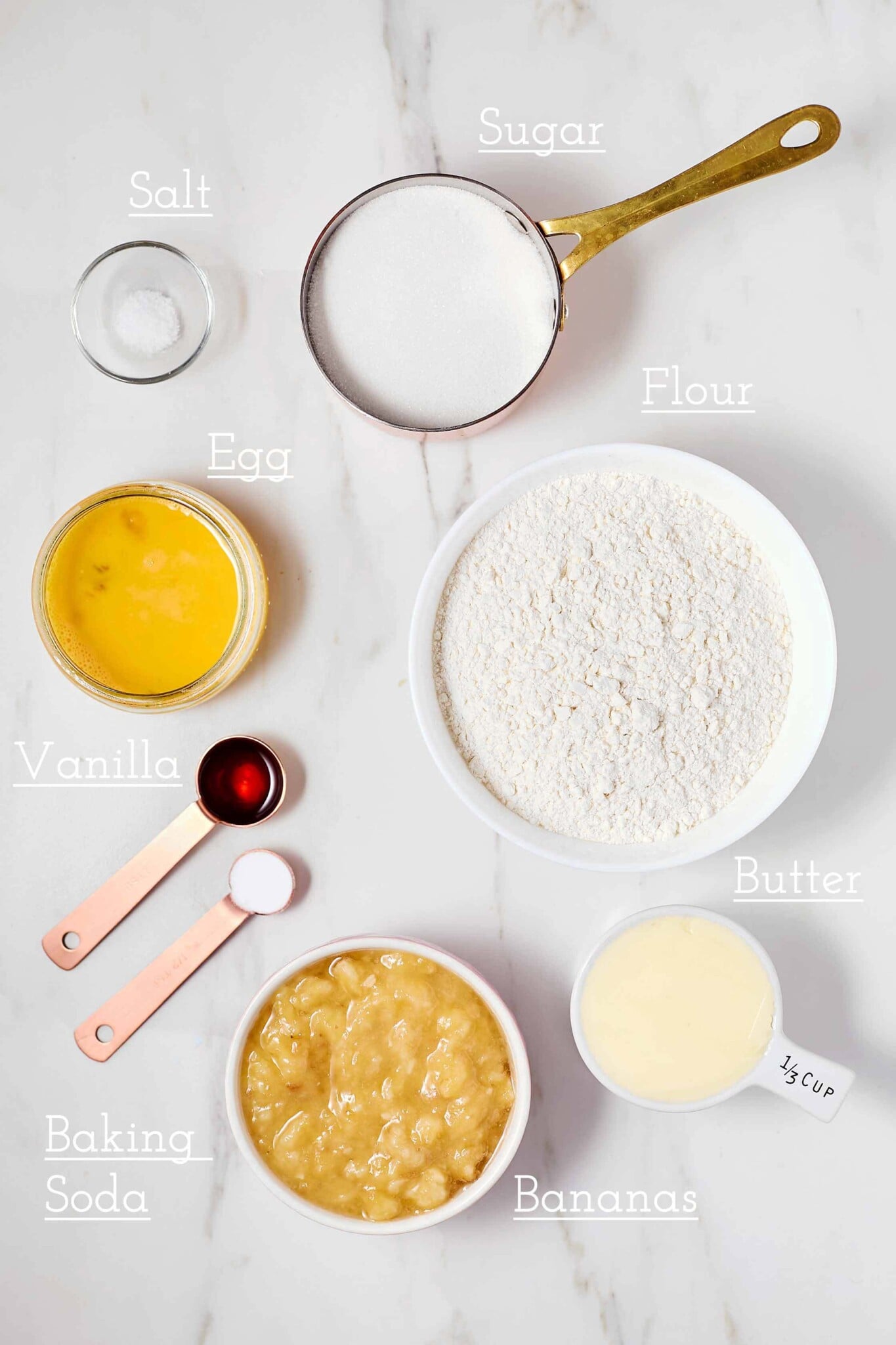 Ingredients for baking laid out on a marble surface, including flour, sugar, salt, eggs, vanilla, butter, baking soda, and bananas, each labeled.