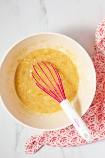 A white bowl containing banana bread batter with a pink whisk on a marble countertop, next to a red and white patterned cloth.