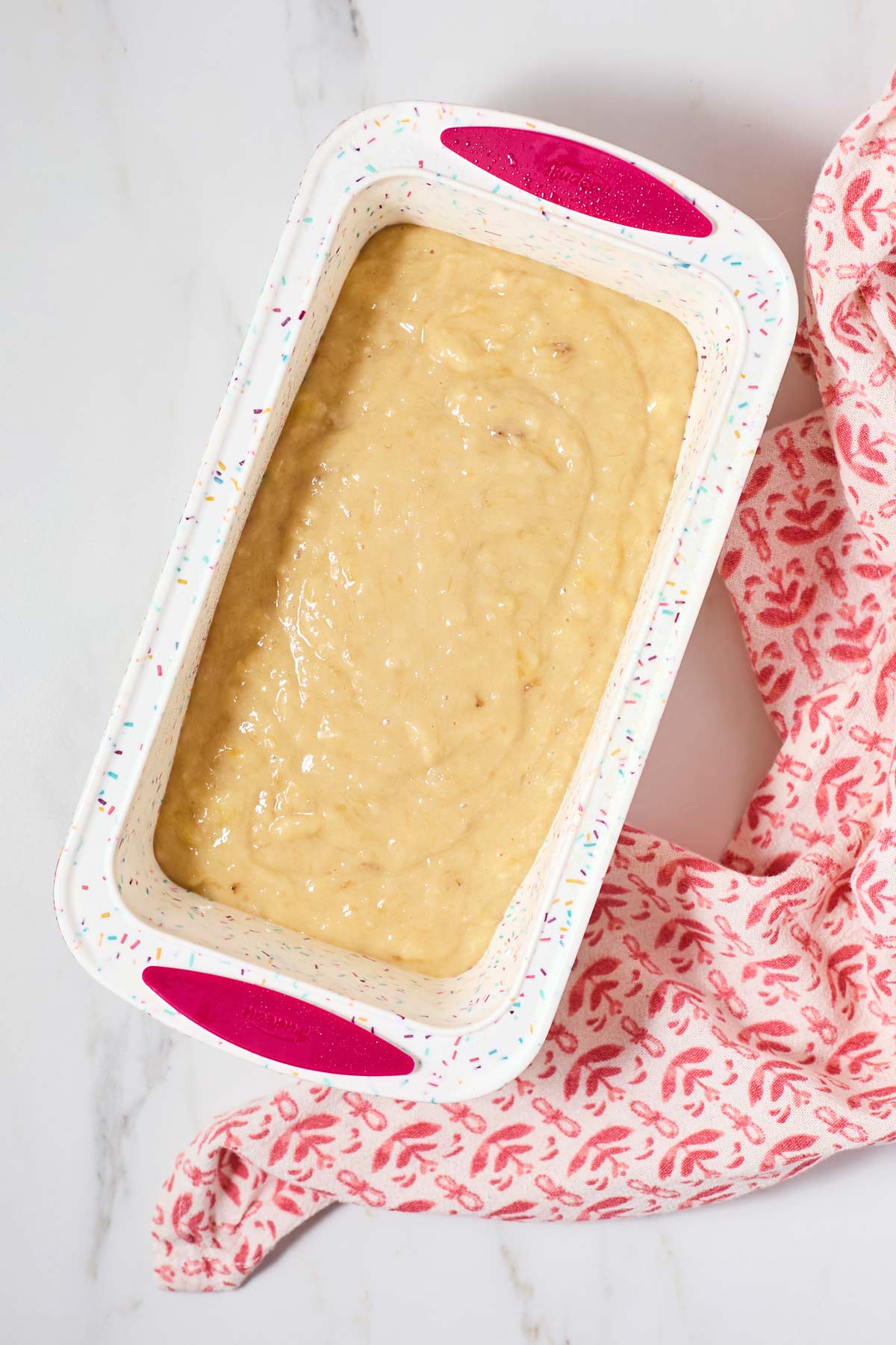 Unbaked banana bread batter in a speckled loaf pan on a marble counter, with a pink patterned oven mitt nearby.