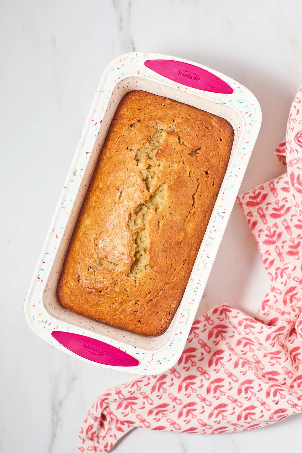 A freshly baked loaf of banana bread in a colorful speckled baking dish, placed on a marble surface next to a red patterned cloth.