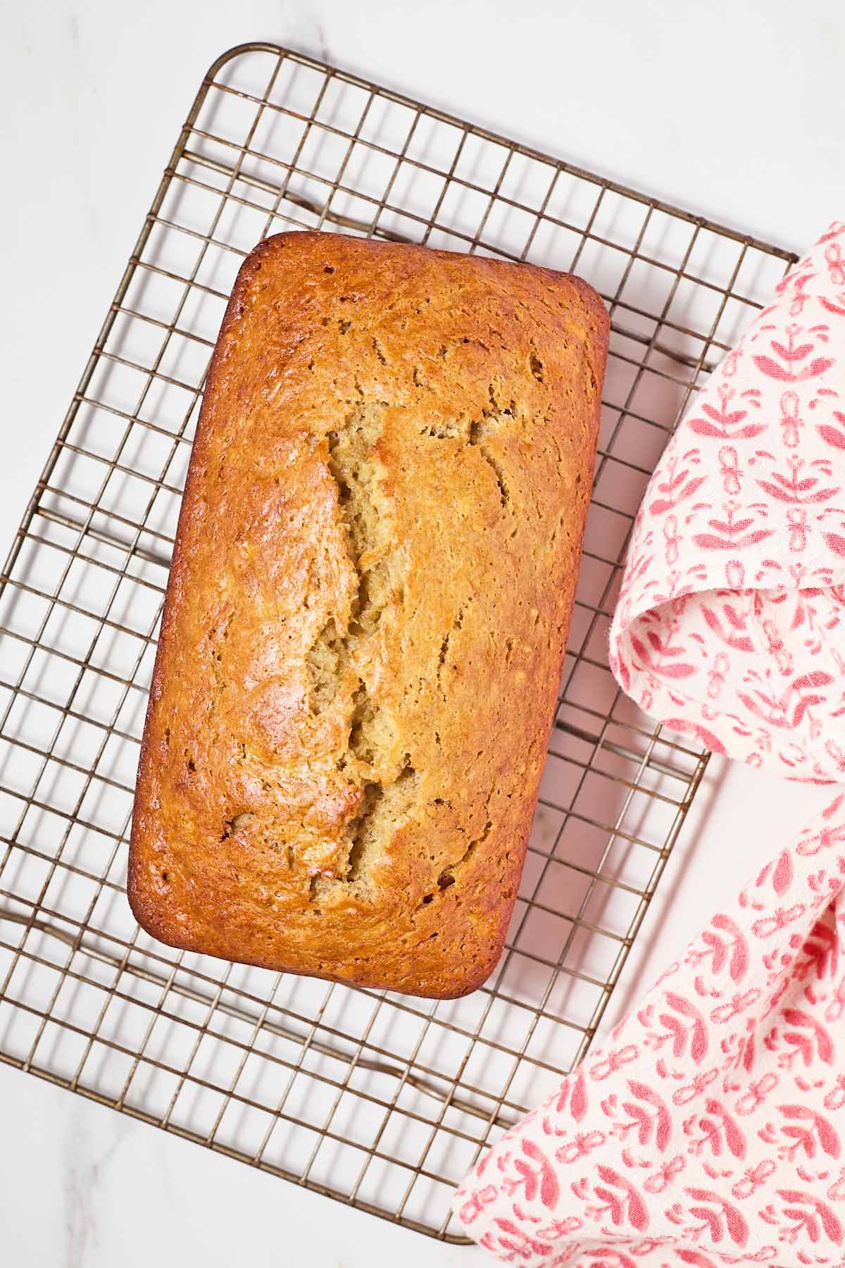 A freshly baked loaf of banana bread cooling on a wire rack, with a pink patterned cloth on the side.