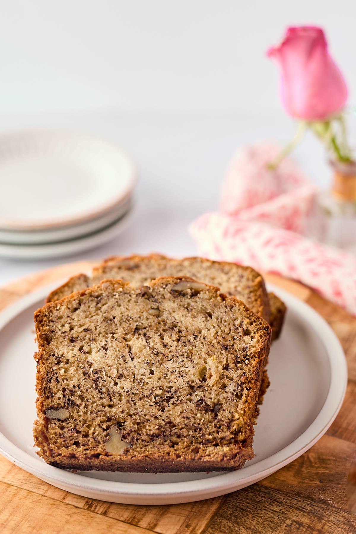 Slices of homemade banana bread on a ceramic plate, with a pink rose and a napkin in the background on a wooden table.