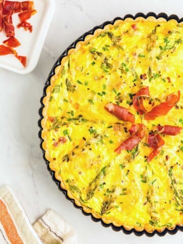 A baked quiche garnished with prosciutto and green herbs on top, placed in a fluted-edge pie dish next to a small square dish with prosciutto slices, and a striped cloth napkin on a marble surface.