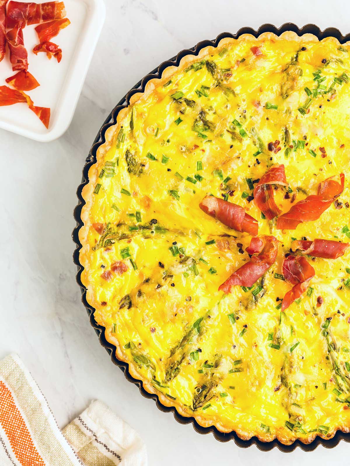 A baked quiche garnished with prosciutto and green herbs on top, placed in a fluted-edge pie dish next to a small square dish with prosciutto slices, and a striped cloth napkin on a marble surface.
