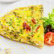 A slice of vegetable frittata garnished with chives and prosciutto, served with a side salad of mixed greens and cherry tomatoes on a white plate with a fork beside it.