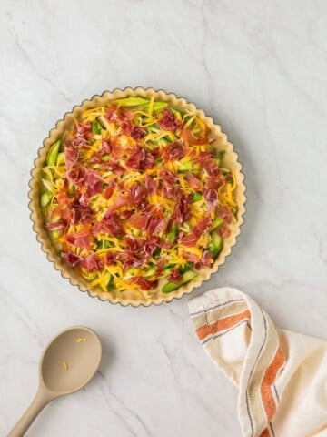 A prepared pie crust filled with shredded zucchini, yellow squash, cheese, and pieces of prosciutto, placed on a marble countertop beside a beige spoon and a striped kitchen towel.