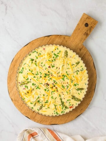 A quiche with visible ingredients such as cheese, chives, and asparagus sits on a round wooden cutting board. A folded striped cloth is in the lower-left corner.