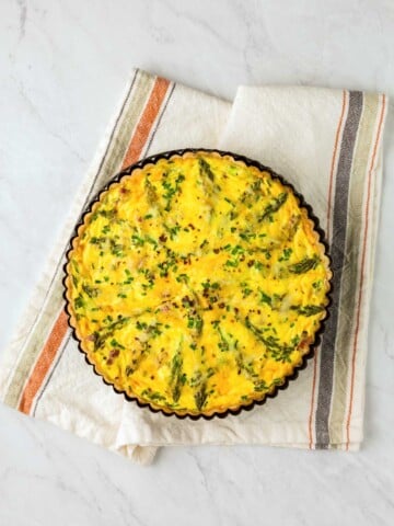 A baked quiche with a golden crust and green vegetable toppings sits on a white and striped dish towel on a marble surface.