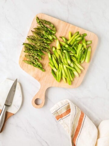 A wooden cutting board with chopped green asparagus and a knife on a marble countertop, next to a kitchen towel.