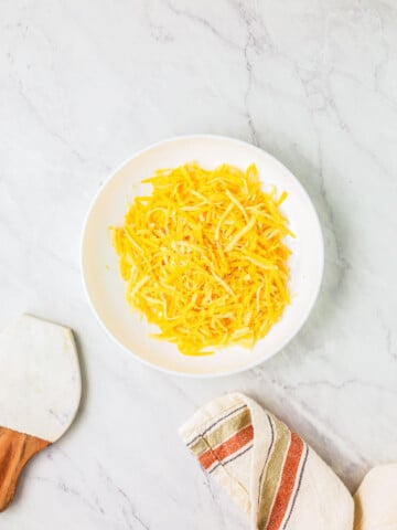 A white bowl filled with shredded yellow cheese placed on a marble surface. A kitchen towel is partially visible beside it.