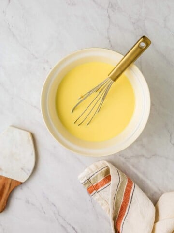 A bowl filled with a quiche batter is on a marble surface with a whisk resting in it. A kitchen towel and a spatula are placed nearby.