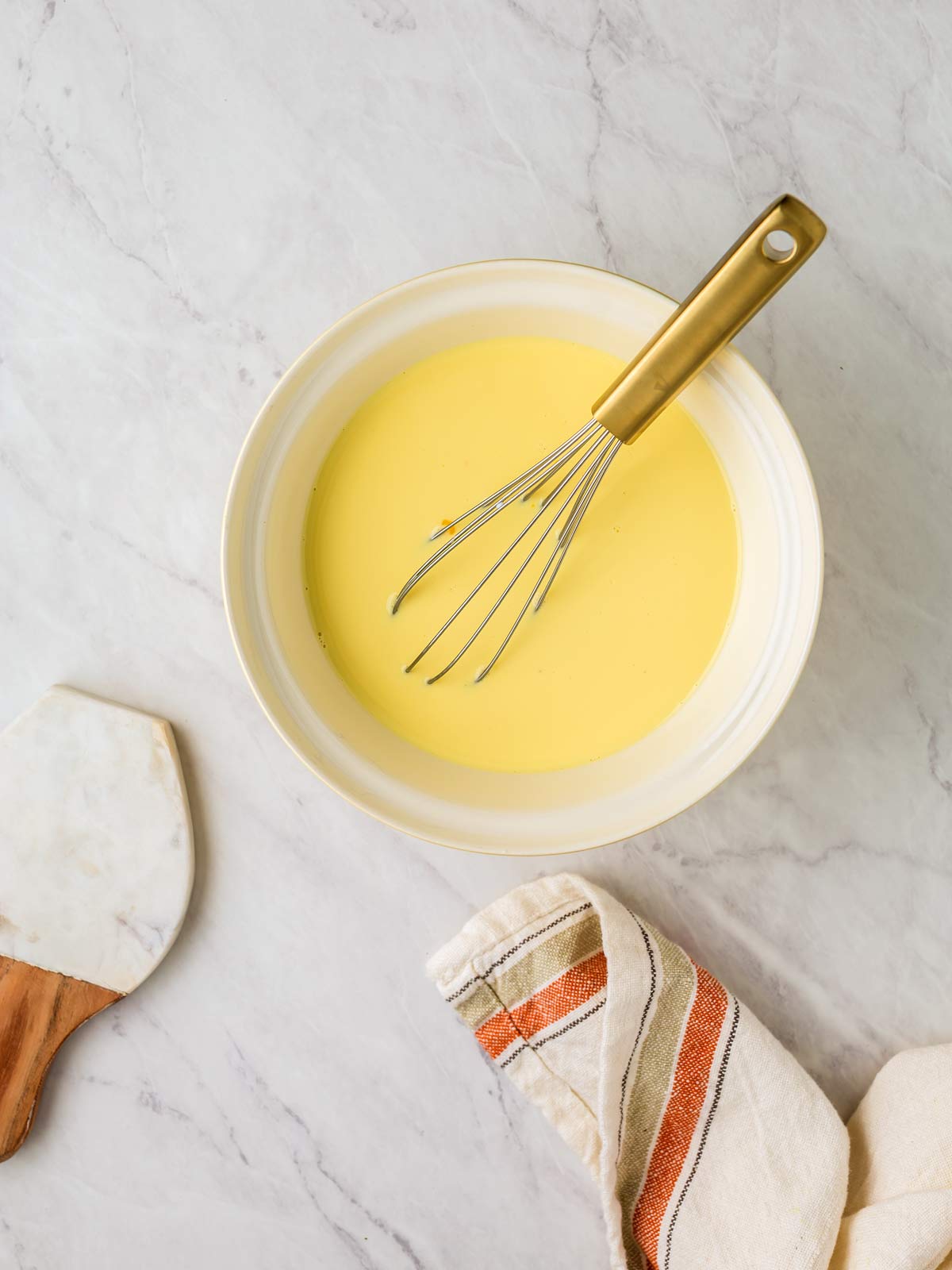 A bowl filled with a quiche batter is on a marble surface with a whisk resting in it. A kitchen towel and a spatula are placed nearby.