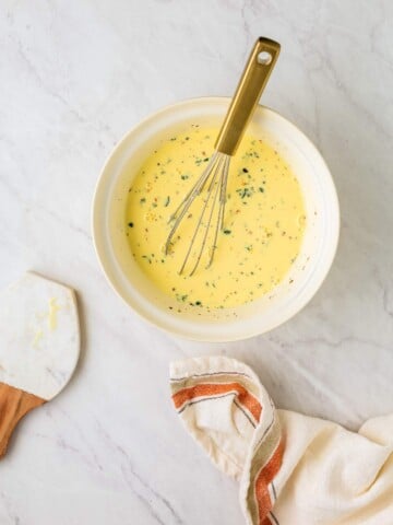 A bowl of seasoned beaten eggs with a whisk, a spatula, and a striped cloth napkin on a light marble surface.