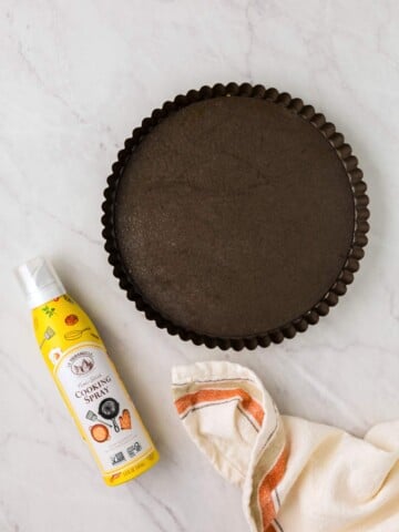 A round, dark-colored fluted pan on a marble surface. Beside it are a can of cooking spray and a folded striped kitchen towel.