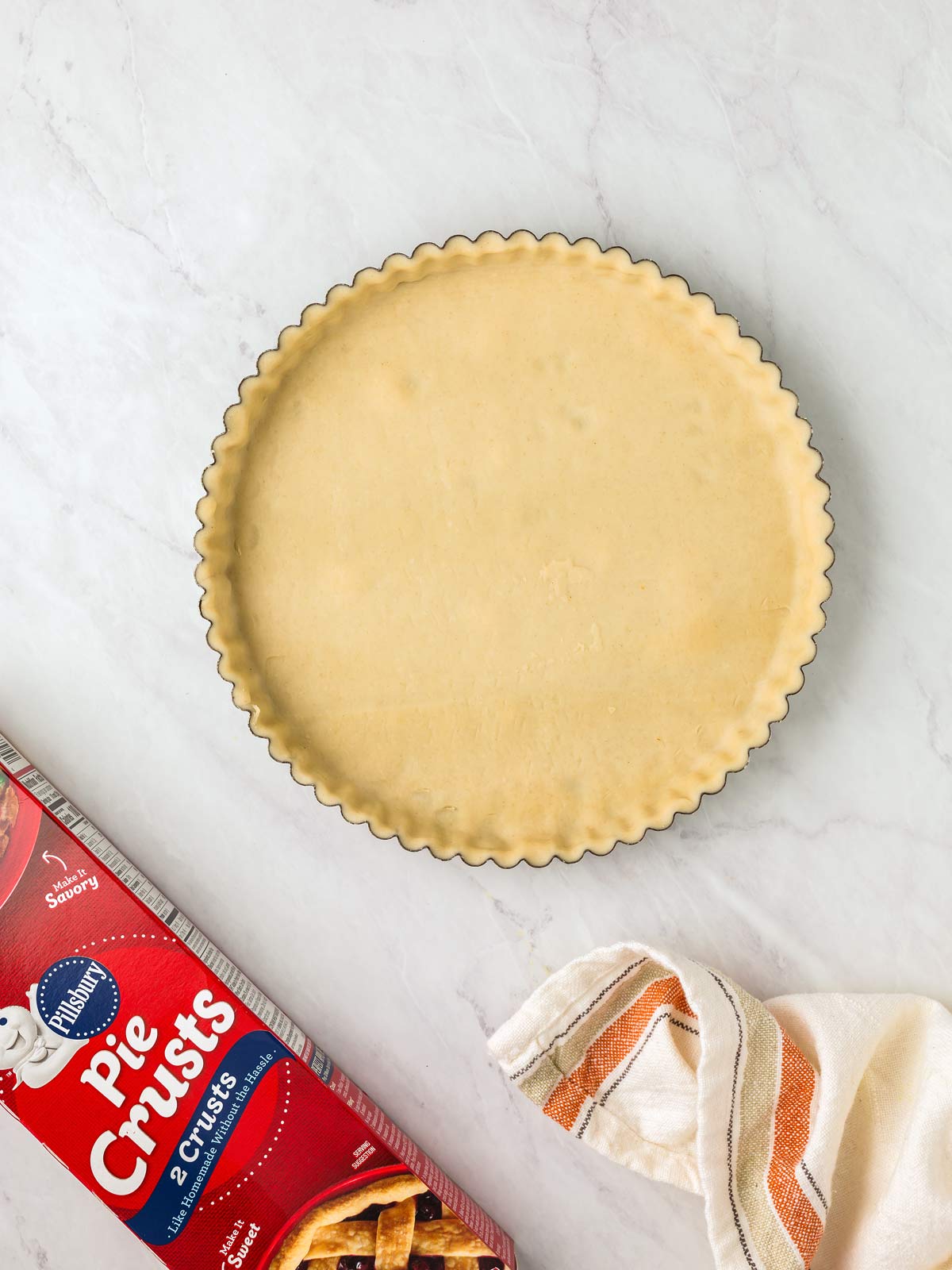 A pie crust in a fluted tart pan is on a white surface near a red box labeled "Pie Crusts" and a striped cloth.