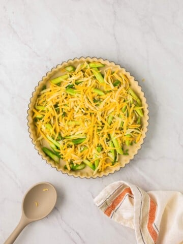 A tart pan filled with uncooked asparagus and shredded cheese next to a beige spoon and a striped cloth on a marble surface.