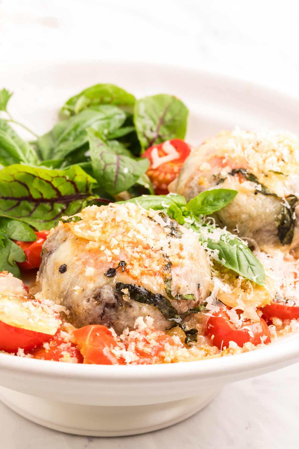 A white bowl contains two mushrooms topped with grated cheese, surrounded by cherry tomatoes and fresh greens.