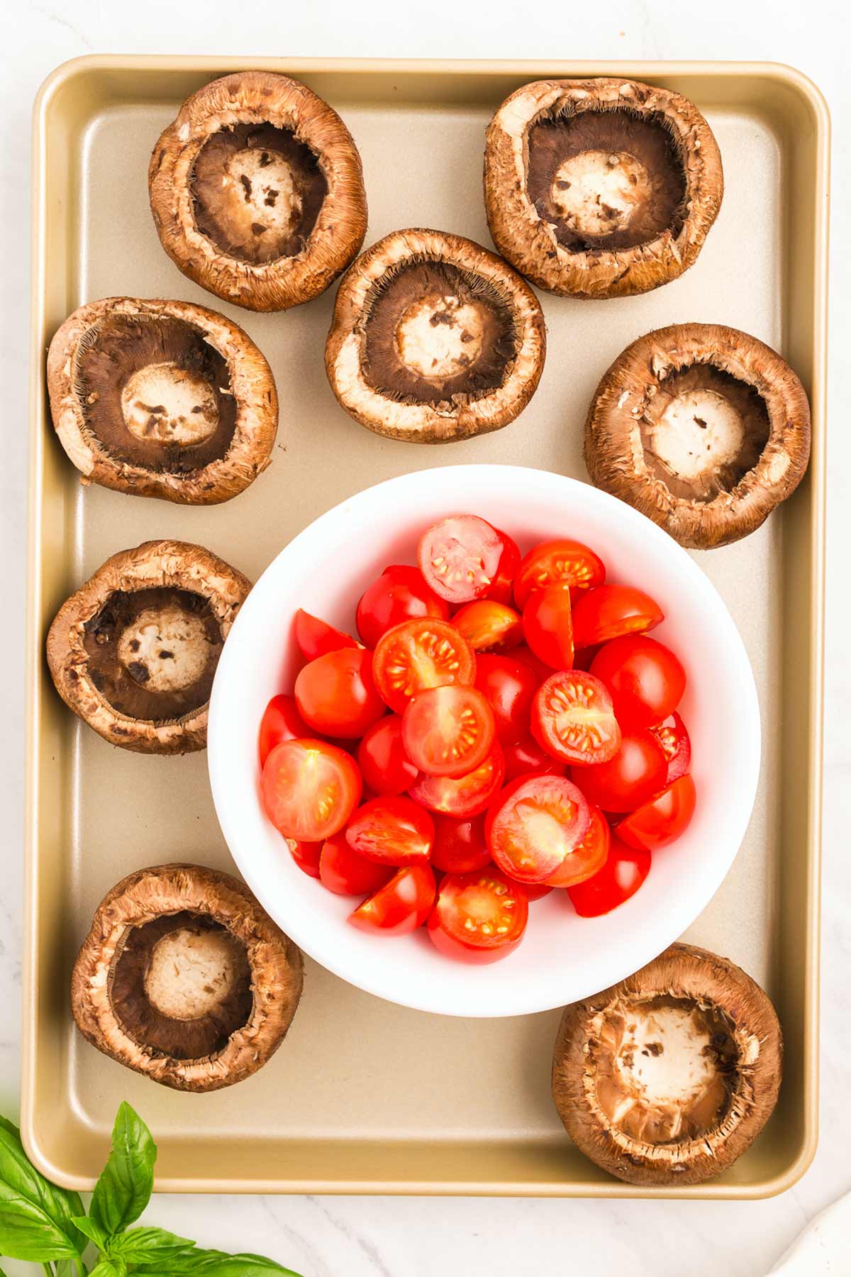 A baking sheet with 8 portobello mushrooms arranged around a white bowl filled with halved cherry tomatoes. Fresh basil leaves are partially visible at the bottom left corner.
