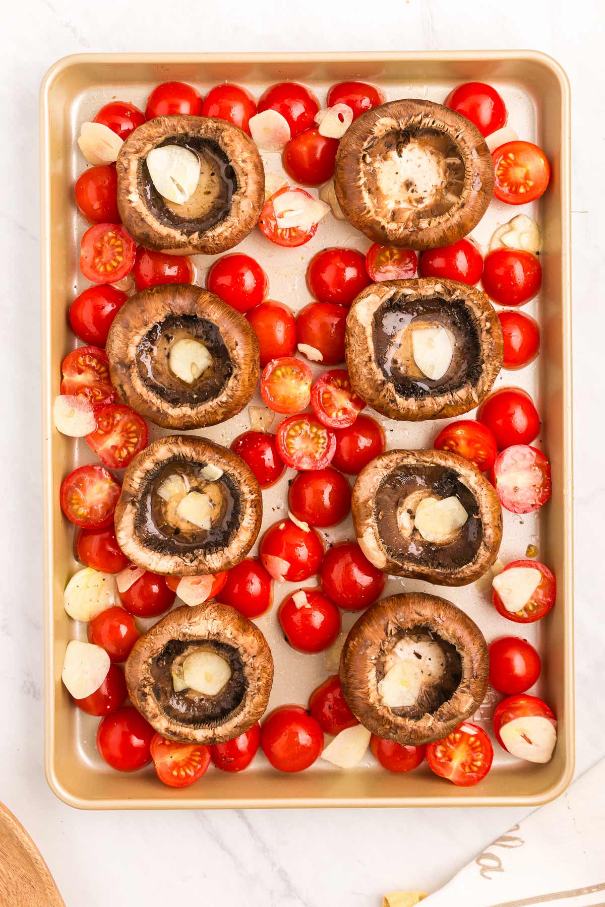 A baking tray with halved cherry tomatoes and large mushroom caps topped with garlic slices, all arranged on a white countertop.