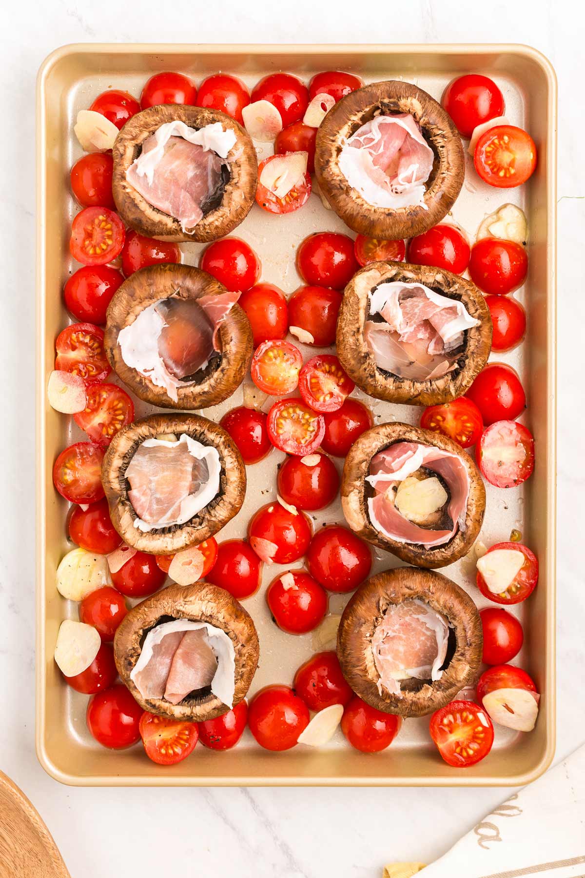 A baking sheet with stuffed mushrooms filled with prosciutto and surrounded by cherry tomatoes and garlic cloves.