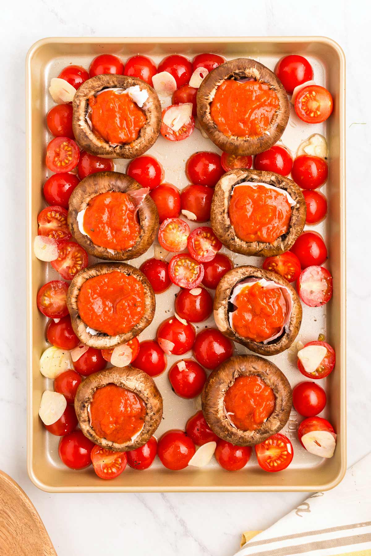 A baking sheet with stuffed mushrooms topped with a red sauce, surrounded by halved cherry tomatoes and garlic slices.