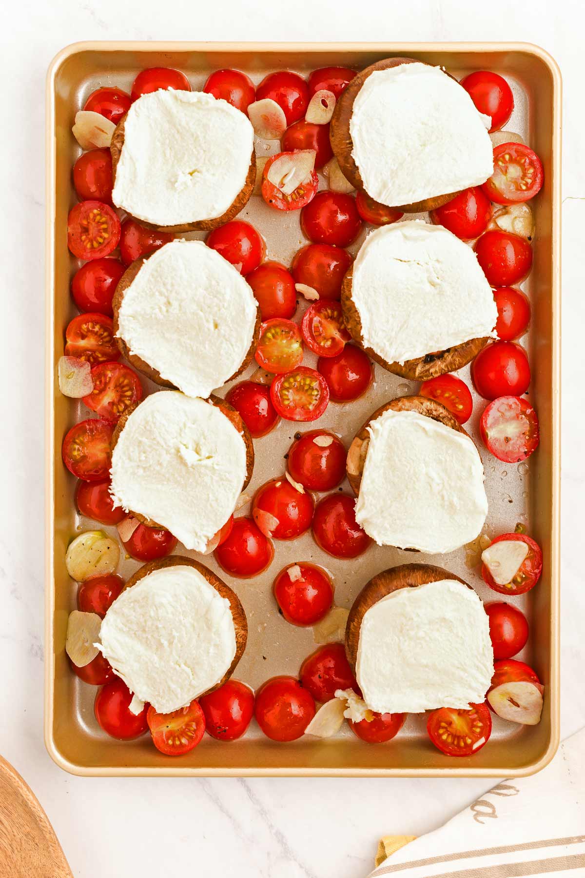 A baking tray with bread slices topped with white cheese and surrounded by cherry tomatoes and garlic slices.