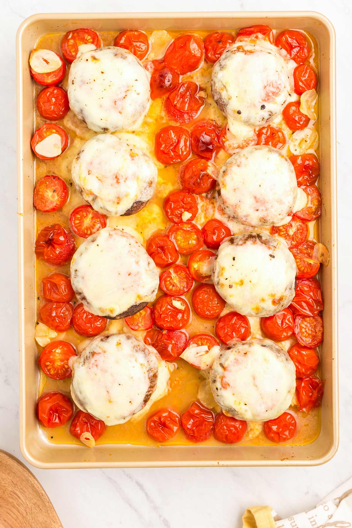 A baking sheet with baked portobello mushrooms topped with melted cheese, surrounded by roasted cherry tomatoes and garlic cloves.