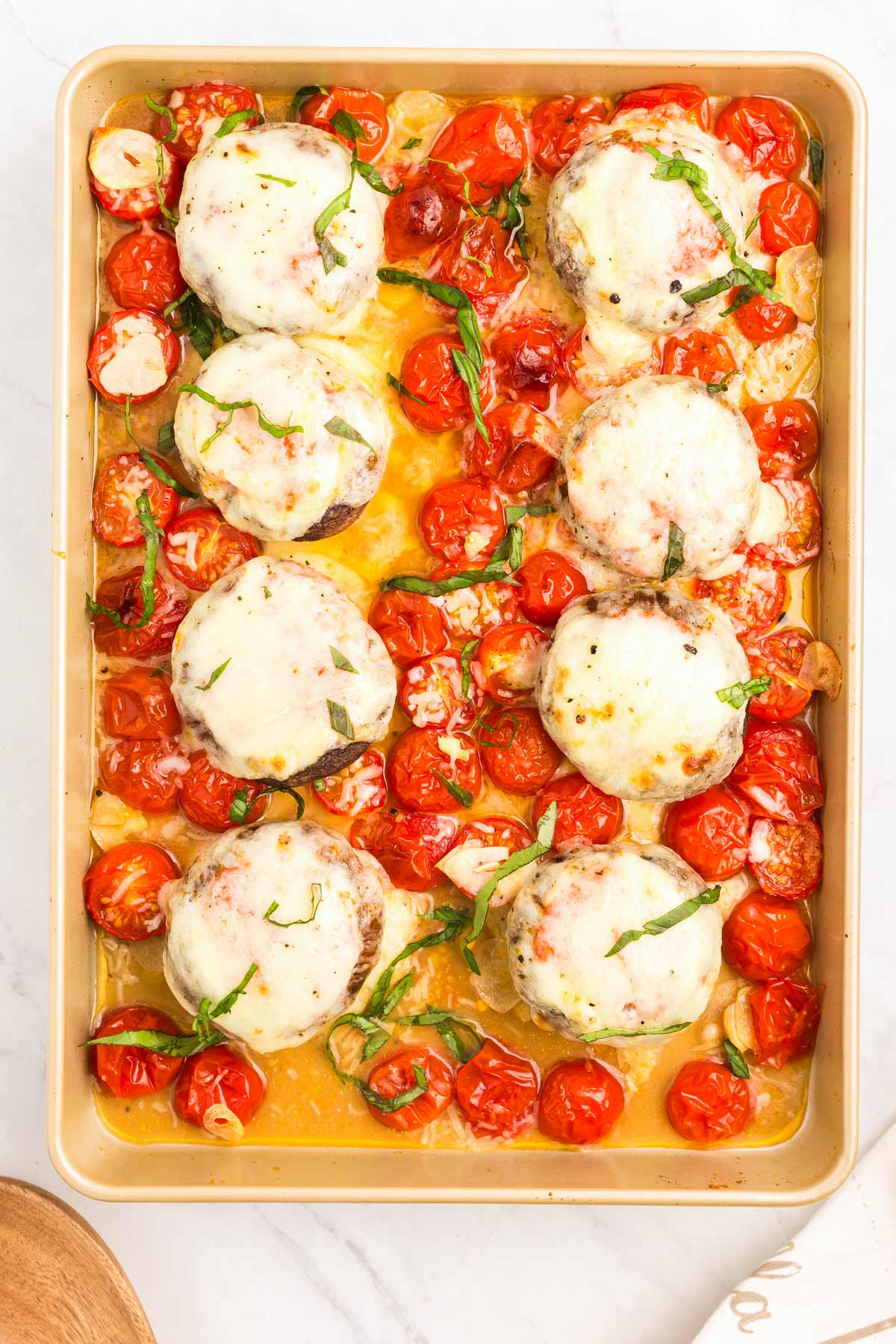Baking tray with stuffed mushrooms topped with melted cheese, surrounded by cooked cherry tomatoes and herbs in a sauce.